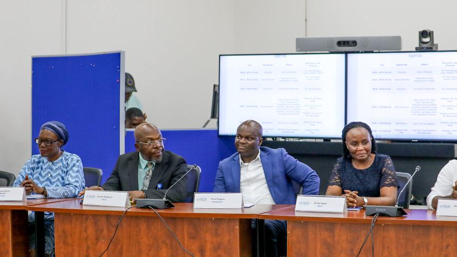 A group of six people all seated at a long desk in a row, each with a nameplate in front of them. Behind them is a banner and two screens. 