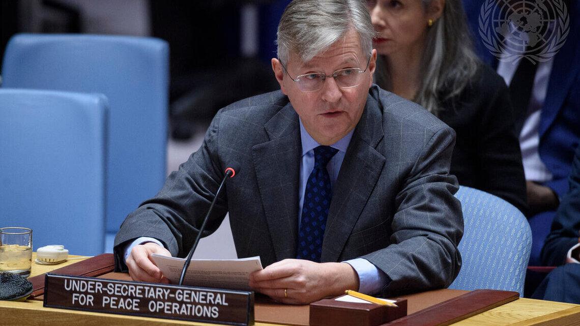 A man in a grey suit sitting a desk behind a sign indicating he is the Under-Secretary-General for Peace Operations. He is speaking into a microphone and addressing the room.