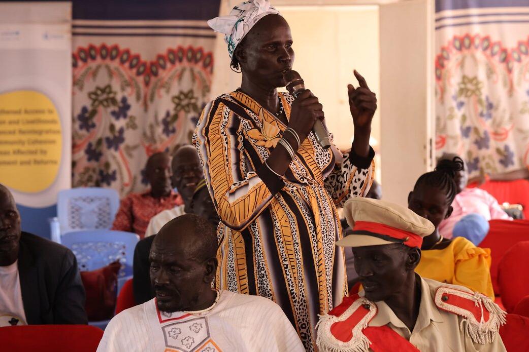 In Kuajok, a harmonization workshop on customary law in Warrap State led to the consensual abolishment of early and forced marriage. Pending on state government approval, this decision will soon be translated into formal law. Person speaking into a microphone while standing among seated participants in a hall decorated with patterned fabric and banners.
