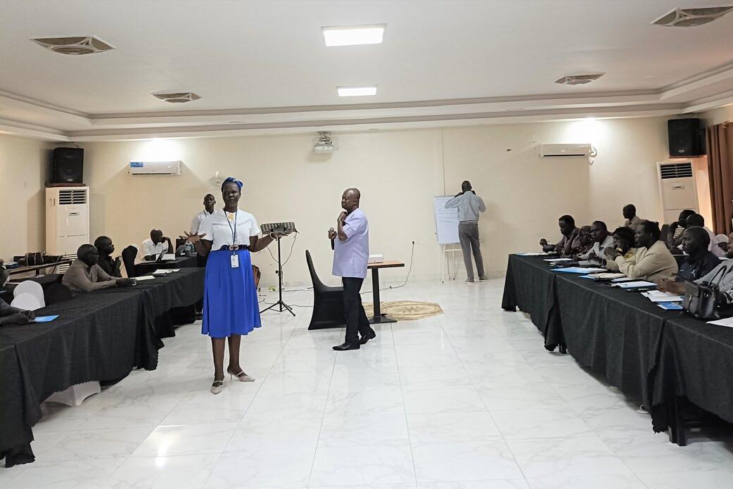 A training session in a conference room with two facilitators standing at the front, participants seated at long tables, and a person writing on a flip chart in the background
