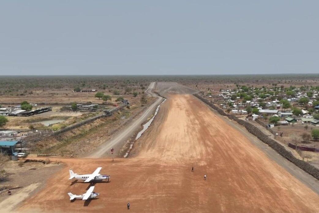 A dirt airstrip with two small white aircraft parked at one end, surrounded by sparse vegetation and scattered buildings under a clear sky in a remote area.