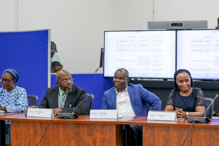 A group of six people all seated at a long desk in a row, each with a nameplate in front of them. Behind them is a banner and two screens. 