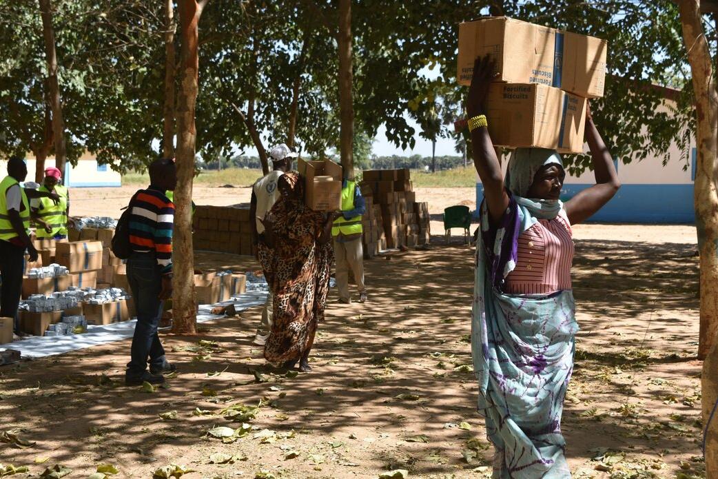 Une femme porte des boîtes sur sa tête, sous des arbres.