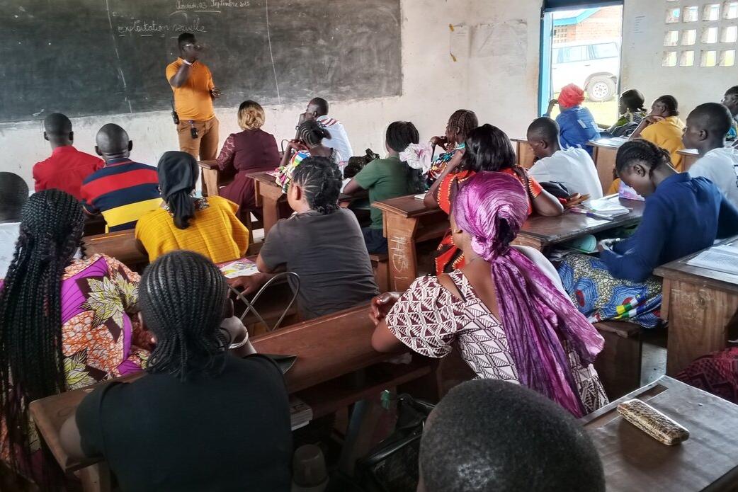 Une salle de classe remplie de gens fait face à une personne debout devant un tableau noir.