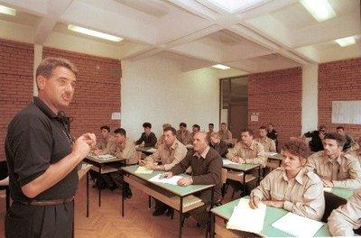 An instructor stands at the front of a classroom addressing a group of trainees seated at desks. The trainees wear matching light-colored uniforms, and the classroom has brick walls and a ceiling with rectangular light panels.