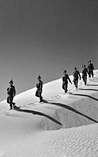 Black and white photo of a group of peacekeepers walking in uniform in single file over a sand dune