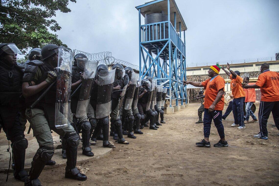 A group of riot police officers holding shields standing in a line with men facing men in orange shirts