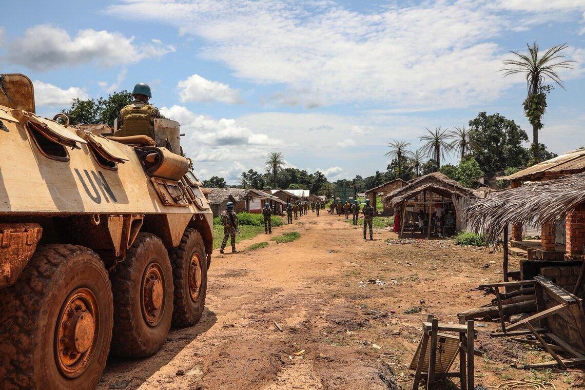 A United Nations (UN) armored vehicle is parked on a dirt road in a rural village. The vehicle is beige with large tires and has "UN" painted on the side. Several soldiers are visible, some standing near the vehicle and others further down the road. The v