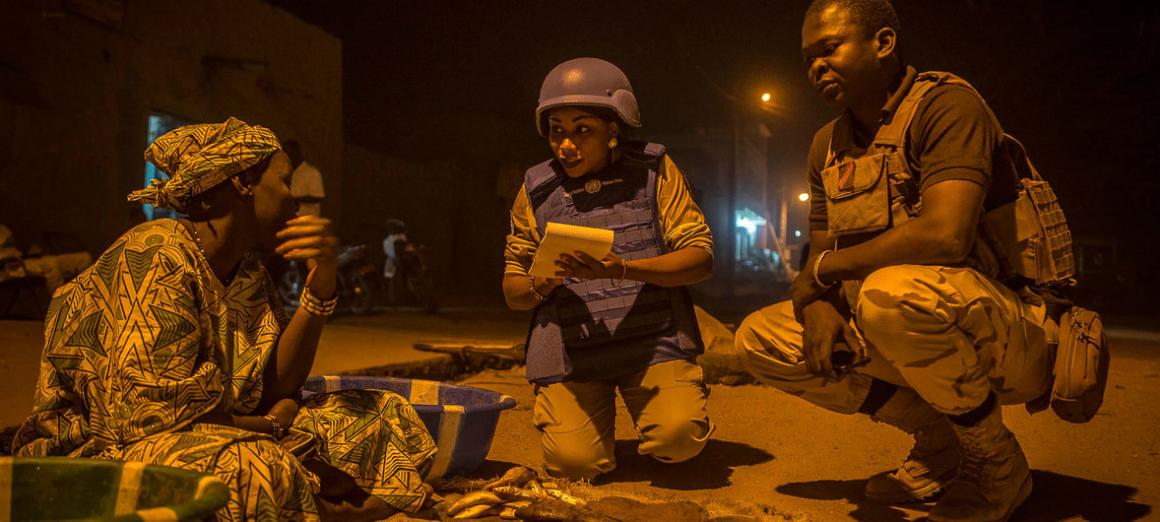 A group of people at a construction site, with one individual pointing and appearing to give instructions to others in uniforms and safety vests, surrounded by metal rebar, dirt piles, and vegetation.