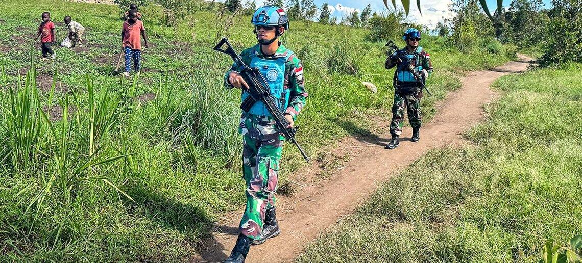 Armed peacekeepers walk along a dirt path, as children look on