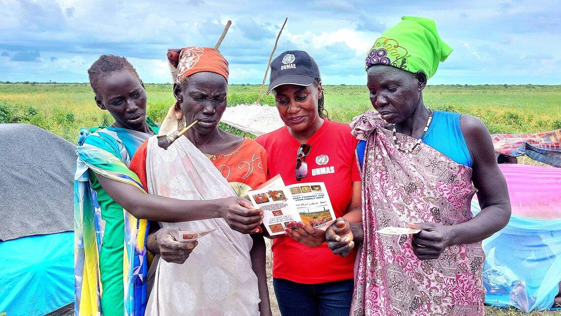 Four women standing together. Three are wearing traditional clothing and the fourth woman is wearing an UNMAS baseball cap and red t-shirt. The four women are looking at UNMAS pamphlets.
