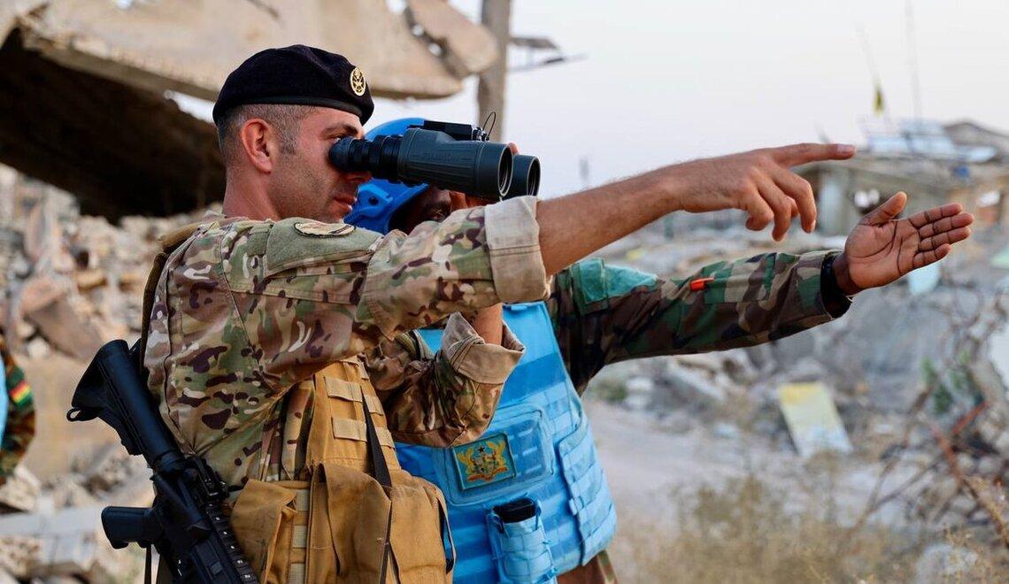 Two military personnel in camouflage uniforms stand amidst rubble and debris in a war-torn area. One holds binoculars while the other points into the distance. Their faces are obscured for privacy. Damaged structures and scattered debris are visible in the background, suggesting a recent conflict or disaster.